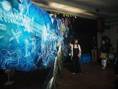 ライトで照らされている青地の上に白色で絵が絵がかれた作品を女性が説明している写真
