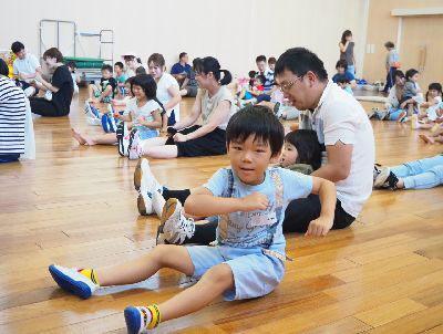 保護者の方と子供たちが床に座っており、そのうちの1人の男の子が両腕を振って上半身を少し左にねじっている写真