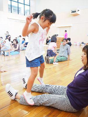 足を広げて座っているお母さんの足の上を跳んでいる女の子の写真