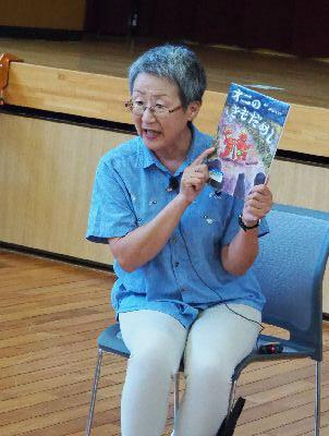 森 一恵さんが椅子に座り「オニのきもだめし」の絵本の表紙を見せている写真