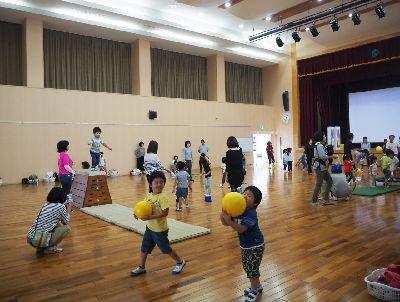 奥ではマット運動や跳び箱で遊んでいる子供たち、手前にはボールを持って歩いている子供たちが写っている写真