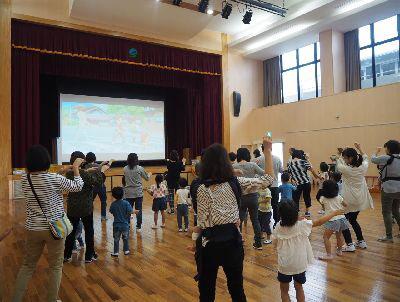 舞台の上に写っているスクリーンを見ながら参加者親子がダンスを踊っている写真