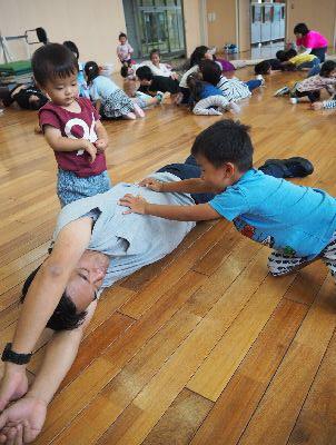 お父さんが腕を頭の上に挙げながら床に寝っ転がっていて、男の子がお父さんの体を両手で押している写真