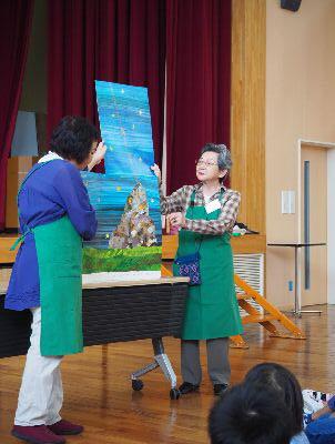 田中さんと長谷川さんが青い背景に岩の絵が描かれた紙芝居を子供たちに見せている写真