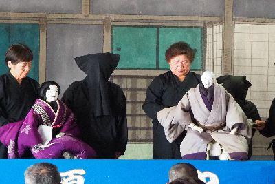 紫色の着物を着た女性の人形の後ろに2人の黒子の人と、グレーの着物を着た男性の人形の後ろに2人の黒子の人が人形を操っている写真
