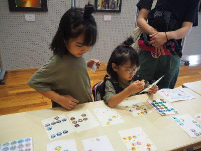 机の上にイラストの描かれたシールが沢山並んで置かれており、左側の女の子はどれにしようかとシールを選んでおり、右側の眼鏡をかけた女の子が出席シールを貼ろうとしている写真