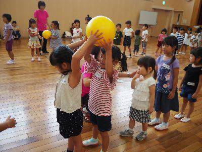 白いティーシャツを着た女の子が後ろにいる赤いボーダーのティーシャツを着た女の子に頭の上で黄色いボールを渡している写真