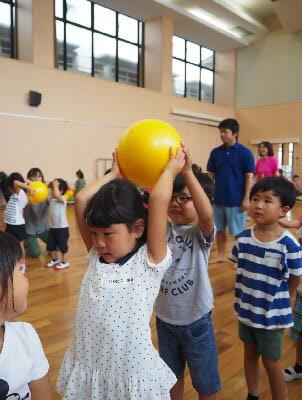子ども達が一列に並んで前を向いており、水玉のワンピースを着た女の子が両手で持った黄色いボールを頭の上から、後ろの眼鏡をかけた男の子に渡して、ボール送りゲームをしている写真