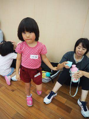 床に座っているお母さんが女の子の水筒の蓋を開けており、水玉のピンクのティーシャツを着た女の子がお母さんの腕を掴んで、カメラ目線で写っている写真