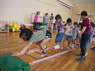 跳び箱にはしごの片方を立てかけて、その上を子ども達が歩いており、先頭の女の子が左手を跳び箱に置いて、右手ではしごの足場を握って四つん這いで昇っている写真