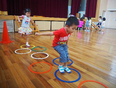 床に輪っかが並べられており、ケンケンパを赤いティーシャツを着た男の子とその後ろには花柄のスカートを着た女の子が挑戦している写真