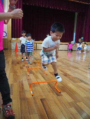 オレンジ色のミニハードルが床の上に3つ置かれており、白いティーシャツを着た男の子が3つ目のミニハードルを飛び越えている写真