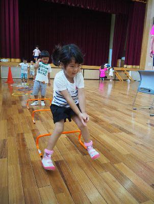 3つのオレンジ色のミニハードルが床に置かれており、白いティーシャツを着た女の子がが3つ目のミニハードルを飛び越え、その後ろで男の子がミニハードルに挑戦しようとしている写真