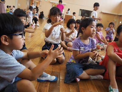 子ども達が集まって床に座っており、左手を握って右手を広げて、手遊びをしている写真