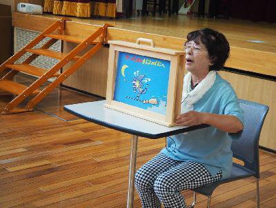 机の上に木枠に入った紙芝居が置かれ、水色のティーシャツを着たスタッフの女性が椅子に座って紙芝居を読んでいる写真