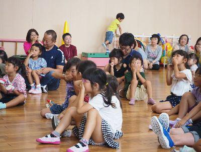 子どもたちが友達や親子で座っており、鼻に手を置いて手遊びをしている写真