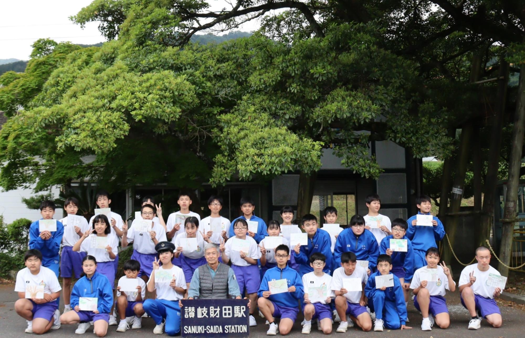 駅舎とタブの木の前で、完成したスケッチ画を手に集合写真