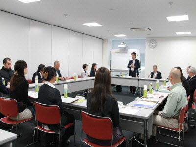 会議室の四角形に並べられたテーブルに参加者たちが座っていて、中央に立っている男性の話を聞いている写真
