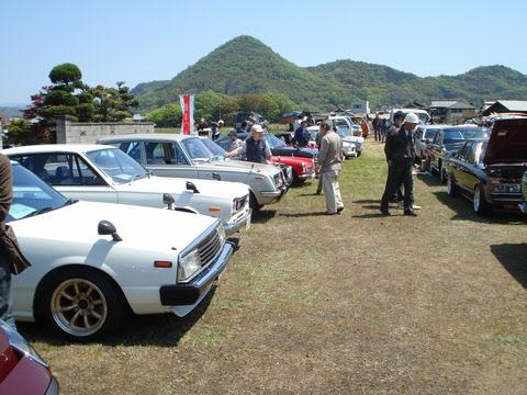 空き地に白や黒のクラシックカーが並んでいて、来場者が車を見学している写真