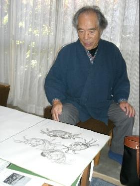 椅子に座りスケッチされた4匹のカニの絵を眺めている西岡 喜三夫さんの写真