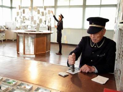 制服を着た中田局長が机の上でスタンプを押しており、奥には8角形の台の上に手紙が展示されており、制服を着た女性の郵便局員の方が手紙を展示している写真