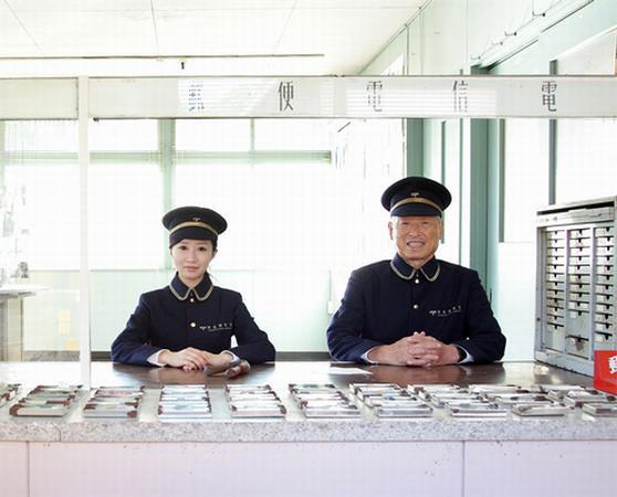 郵便局員の帽子を被り、制服を着た年配の男性と若い女性が並んで座っている写真