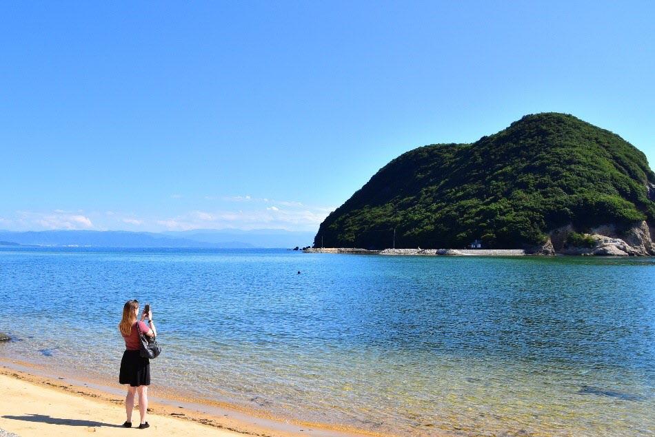 透き通った海の奥にある丸山島の写真を撮る女性の写真
