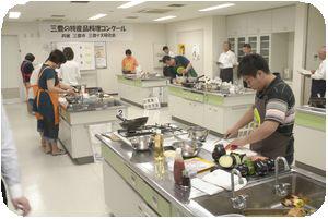 参加者達がそれぞれに調理台で料理をしている所を審査員の方々が見ている写真