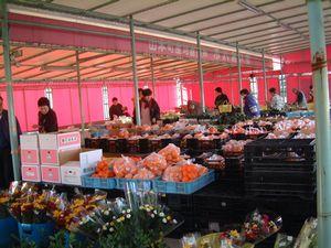屋外テントの中に果物や花などが並び、多くの買い物客が商品を見ている写真