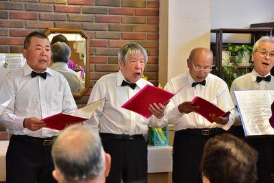 ミントクラブのメンバー4人の男性が赤いファイルを手に持ち指揮を見ながら歌を歌っている写真