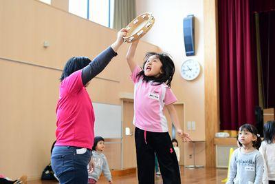 スタッフが高く上げたタンバリンを女の子が叩こうと手を伸ばしている写真