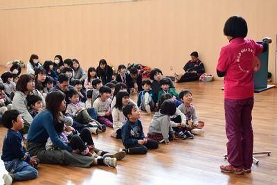 スタッフの女性が座っている子供達の前で紙芝居を読んでいる写真