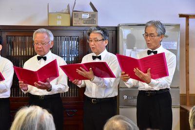 3人のミントクラブの男性が手に持った赤いファイルを見ながら真剣に歌を歌っている写真