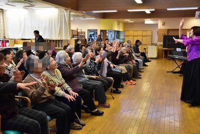 豊岡 真由美先生が観客たちの前で右手を額の前に左手を胸の高さに挙げているポーズをとっており、観客たちも豊岡 真由美先生と同じポーズをしている写真