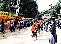 境内の前の端に屋台が並び、中央を衣装を着た子供たちが行列で歩き、袴を着た大人たちが立っている写真