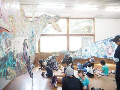 大きな鯨の作品が飾られた部屋で、子供達が座って作品を作っている写真