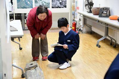 座ってノートを取っている男子生徒と男子生徒を覗き込んでいる年配の女性の写真