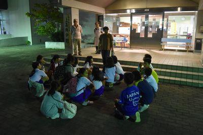 夜、子供達が建物の外で座って女性のお話を聞いている写真