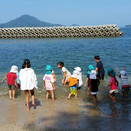 裸足になって波打ち際で遊んでいる園児と小学生の写真