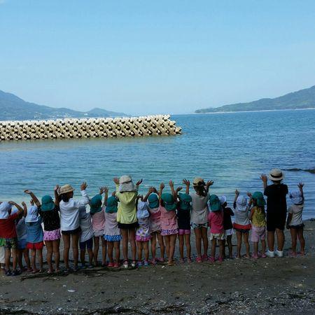 砂浜で海に向かって横一列に並んで両手をあげている園児と小学生の写真