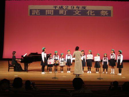 緑のベレーボーをかぶり白のブラウスに黒のスカートでステージに並び合唱を披露している子供たちと先生達の写真