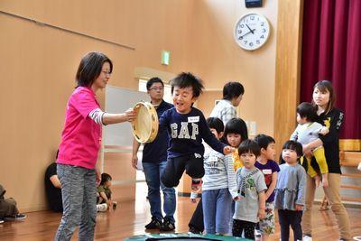トランポリンを跳んで横に立っているスタッフが持っているタンバリンをタッチしようとしている男の子の写真