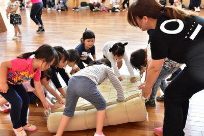 沢山の子供たちと一人の女性が体育マットを持ち上げようとしている写真