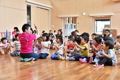 床に座っている子供たちが前に座っているスタッフの真似をして人差し指を立てて両手を挙げている写真