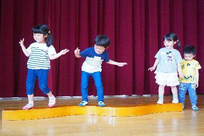 曲線平均台が床に置かれており、ストライブのティーシャツの女の子とジーパンを履いた男の子が手でハサミを作ってカニさんになって横歩きで平均台の上を歩いている写真