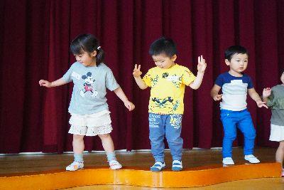曲線平均台が床に置かれており、イラストの描かれたティーシャツを着た女の子が下を見ながら横歩きをしており、隣の黄色いティーシャツを着た男の子が手でハサミを作ってカニさんになって横歩きで平均台の上を歩いている写真