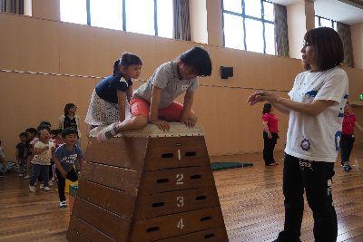 6段の高い跳び箱の上にオレンジ色のズボンをはいた女の子が両手をついてまたがっており、側にはスタッフの女性が見守っている写真