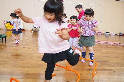 床に置かれたオレンジ色のミニハードルを片足を挙げてピンク色のティーシャツを着た女の子が飛び越えている写真