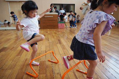 床に置かれたオレンジ色のミニハードルを紫色のティーシャツを着た女の子が2つ目を飛び越えており、後ろに続くピンクのティーシャツを着た女の子が右足を挙げて飛び越えようとしている写真