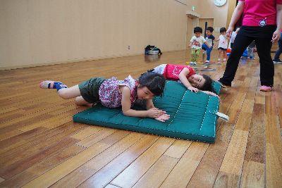 緑色の体育マットの下に物が置かれ、凹凸のあるマットの上を子ども2人が寝転んで両手を上で重ねてコロコロと体育マットの上を転がっている写真